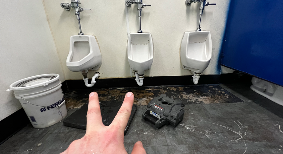 Evan from Larry's Plumbing pointing at two broken urinals in the men's restroom at Sports World Athletics in Rockwall, Texas.